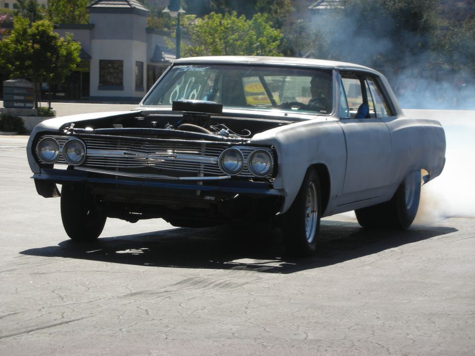 Black-and-primer 1960s muscle car doing a smoky burnout with blower poking through hood at a car show