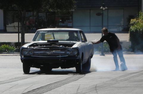 1998 Ford Crown Victoria - man leans on the trunk of a black-and-primer muscle car steadying it as the rear tires smoke in a burnout.