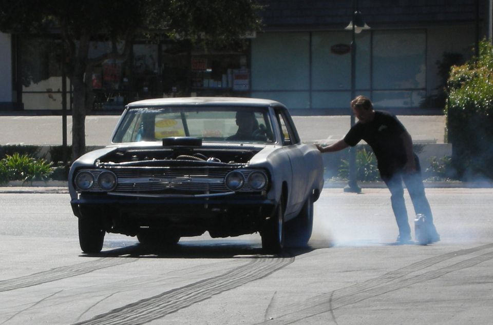 Man steadies a black-and-primer muscle car as it smokes its rear tires in a burnout outdoors