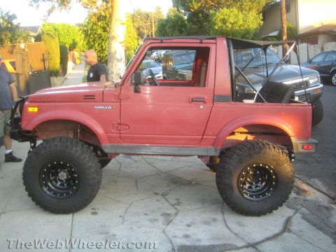 1987 Suzuki Samurai - This red 1987 Suzuki Samurai features prominent off-road tires and a roll cage, seen parked on a residential street.