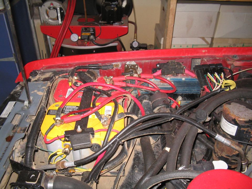 Dual batteries, red cables and a blue ignition module in the engine bay of a red vehicle in a garage