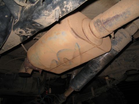 1989 Ford Bronco - The undercarriage of a vehicle shows a dusty exhaust muffler and driveshaft, with leaf springs and a shock absorber also visible.