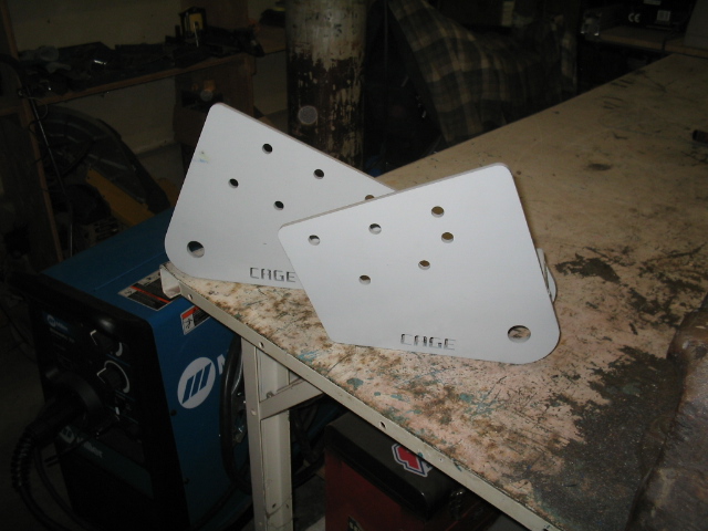 Two flat steel CAGE-stamped suspension link brackets with multiple bolt holes on a workbench