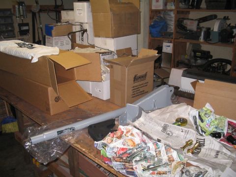 1989 Ford Bronco - A gray suspension component, possibly for a vehicle's chassis, lies on a cluttered workbench alongside boxes and hardware.