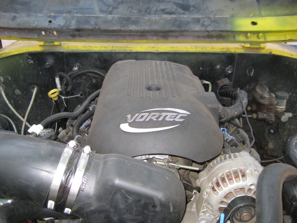 Vortec engine cover with air intake tube and alternator visible. Supermotors.net logo at bottom right