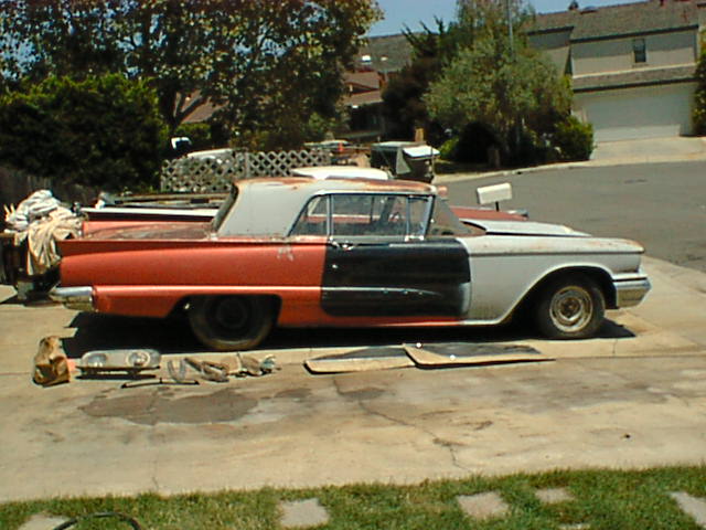 Side view of a weathered orange 1959 Thunderbird with black door and primered rear, parts laid on driveway
