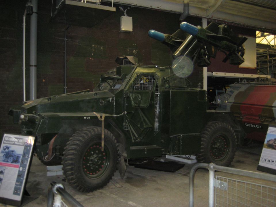 Green armored vehicle with missile launcher on top, parked indoors. Another vehicle with camouflage is visible to the right