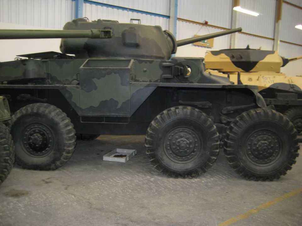 Camouflaged armored car with six wheels and a turret, parked indoors next to another vehicle