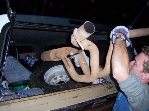 1991 Ford Bronco - A person removes an exhaust manifold from the cargo area of a vehicle, with a spare tire visible.