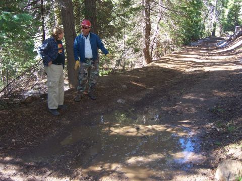 1991 Ford Bronco - Two men converse on a dirt trail in a wooded area, with a muddy puddle reflecting the sky.