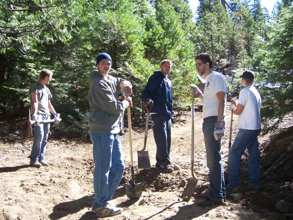 Five men with shovels and tools stand on a dirt path in a wooded area, appearing to be doing trail maintenance