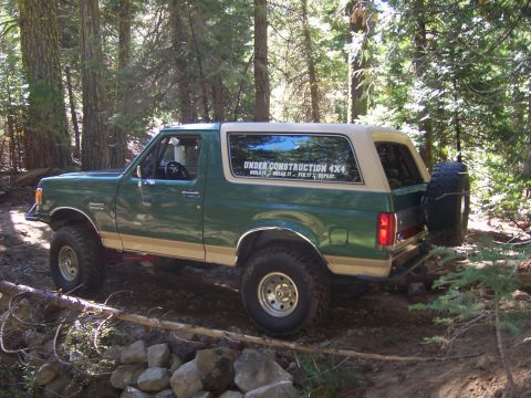 1991 Ford Bronco - A green 1991 Ford Bronco with a tan removable top is shown on a wooded trail, a spare tire mounted on the rear.