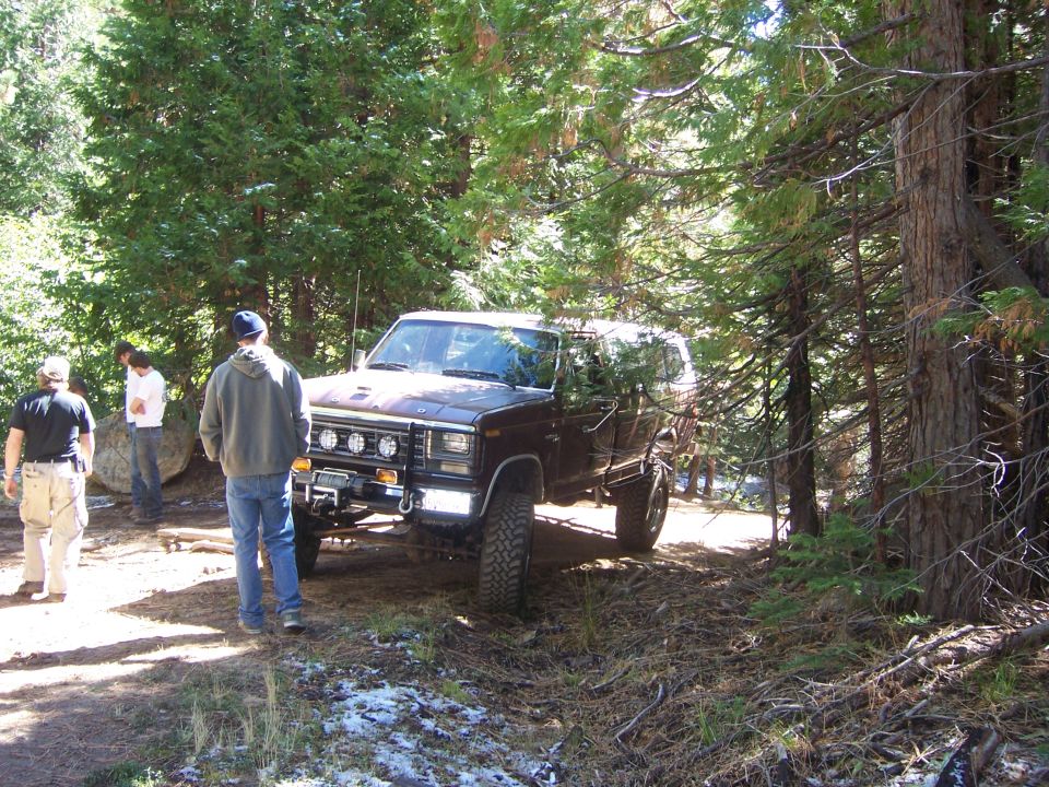 Brown Ford Bronco with winch and auxiliary lights parked on a dirt path in a forest, people standing nearby