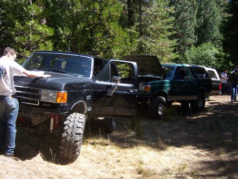 1991 Ford Bronco - Several Ford Broncos are parked off-road in a wooded area, with some people gathered around.