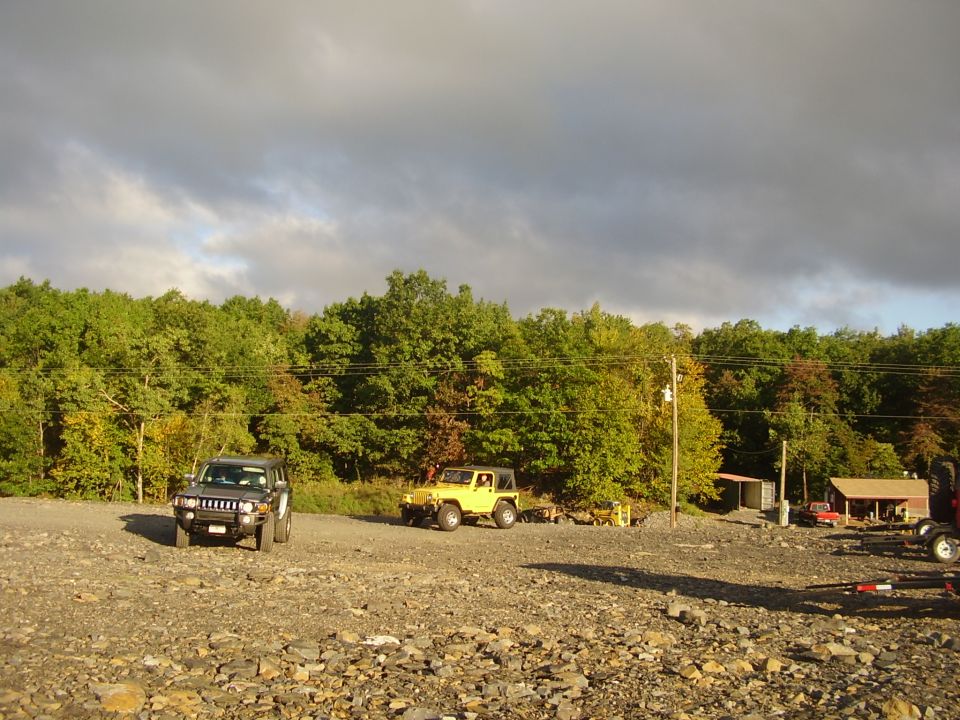 Black Hummer H3 and yellow Jeep Wrangler parked in a gravel lot with trees in the background
