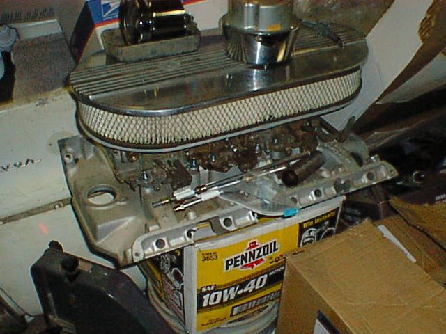 Intake manifold with carburetors and chrome oval air cleaner resting on a Pennzoil 10W-40 box in a garage