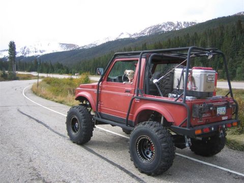 1987 Suzuki Samurai - A modified red 1987 Suzuki Samurai with a dog inside is parked on a scenic mountain road.