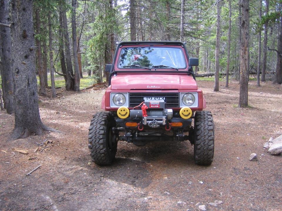 1987 Suzuki Samurai parked in a forest, front view, with a winch and yellow smiley face fog lights