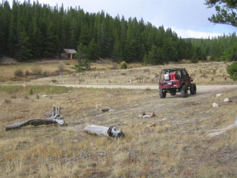 1987 Suzuki Samurai - A red off-road vehicle is parked on a dirt road with a wooded area and a small cabin in the background.
