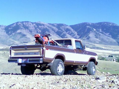 1979 Ford F-150 - A two-tone brown and white 1979 Ford F-150 pickup truck is parked on a rocky hill, carrying a dirt bike in its bed with mountains in the ba…