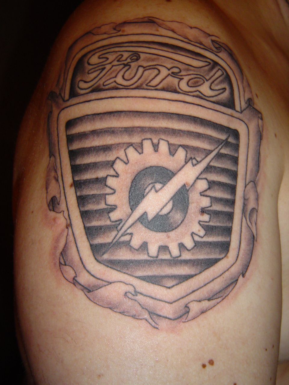 Upper-arm tattoo of a 1950s Ford truck hood emblem with Ford script, gear and lightning bolt