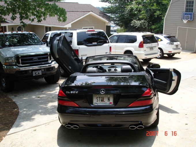 Rear of black Mercedes SL500 convertible, top down, trunk open, in a driveway with other vehicles.