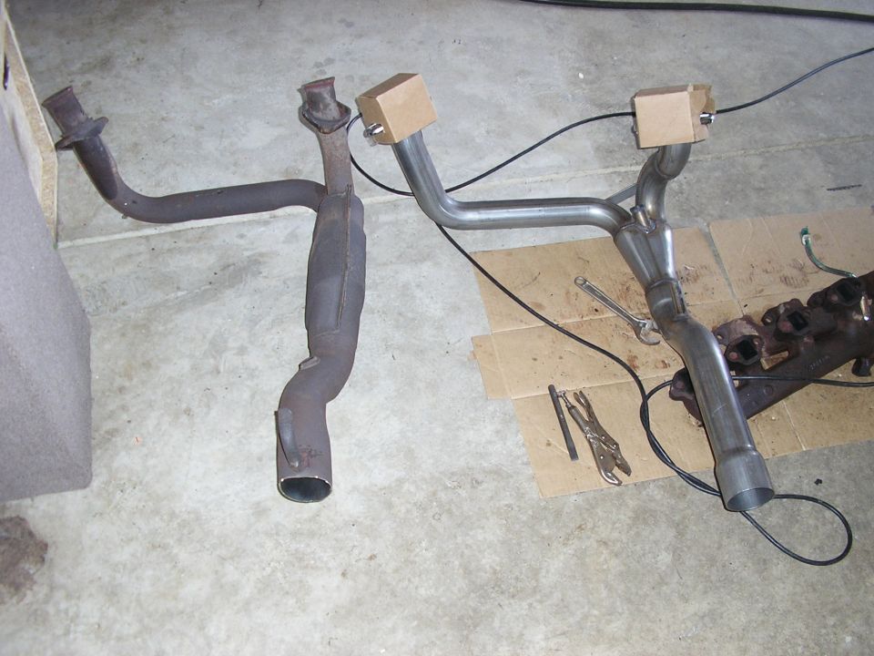 Rusty stock exhaust Y-pipe beside a new stainless replacement Y-pipe on a garage floor