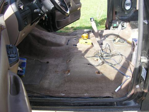 1996 Ford Bronco - Interior of a Ford truck with the front seats removed, shot through the open driver's door onto the brown carpet with exposed seat-mount bo…