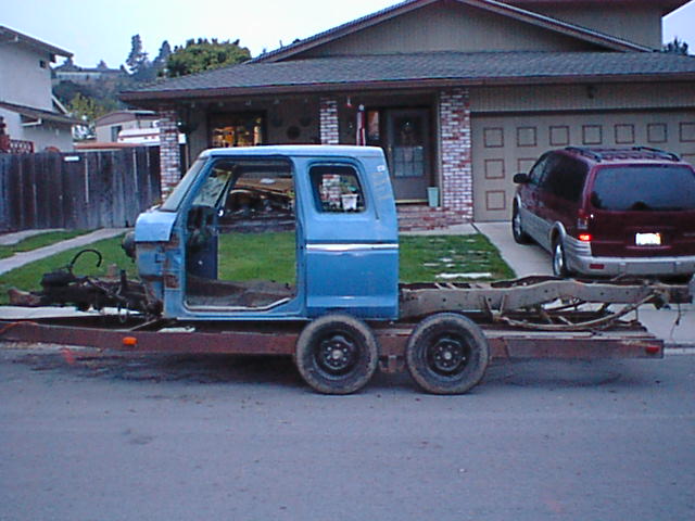 Blue Ford truck cab on bare frame, no doors or bed, sitting on a tandem-axle trailer in front of a house