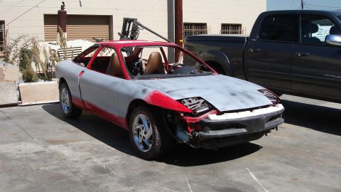 1996 Ford Probe - A sports car in primer and red accents sits next to a pickup truck, awaiting restoration.