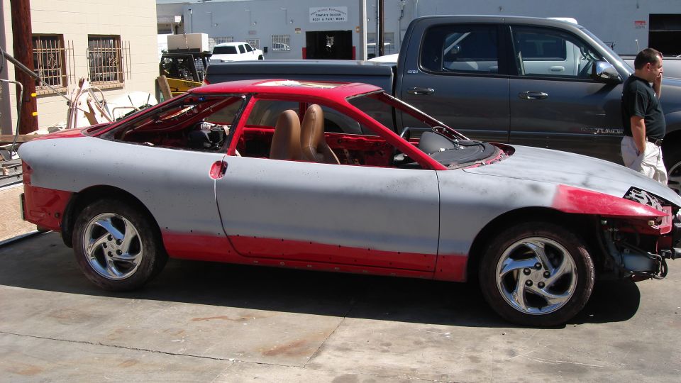Primered and red car with roll cage, tan seats, and chrome wheels, side view