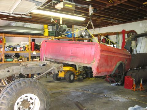 1990 Ford Bronco - A red truck bed is suspended by a hoist in a garage, with the vehicle's chassis and a large tire visible below.