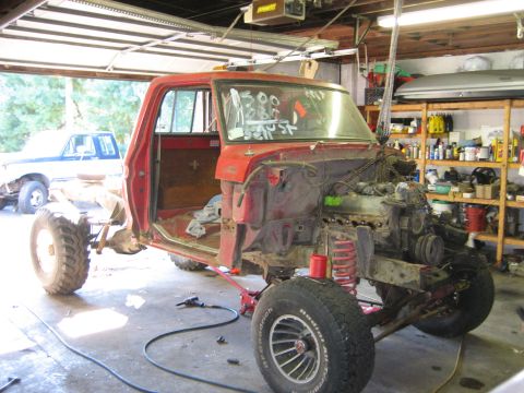 1990 Ford Bronco - This red truck chassis, missing its body panels, is undergoing work in a garage. A second truck is partially visible in the background.