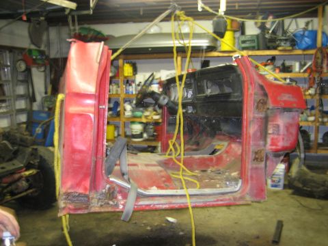 1990 Ford Bronco - A red truck cab, likely from the late 1970s, is suspended by a hoist in a garage, revealing its interior and significant rust.