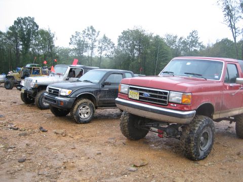 1990 Ford Bronco - Several modified trucks and SUVs are parked on a dirt track with trees in the background.