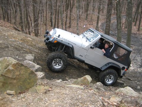 1990 Ford Bronco - A silver Jeep Wrangler navigates a challenging rocky trail in a wooded area.