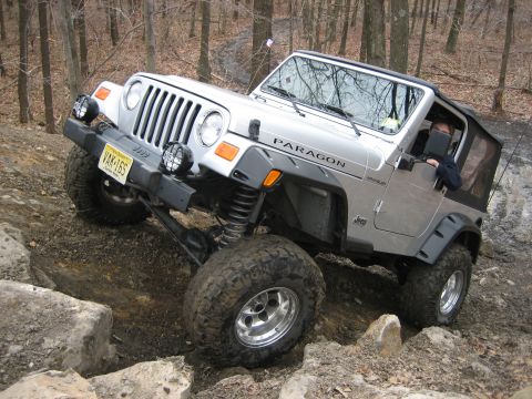 1990 Ford Bronco - This silver Jeep Wrangler is shown tackling a challenging rocky terrain, with the word 'PARAGON' visible on its side.