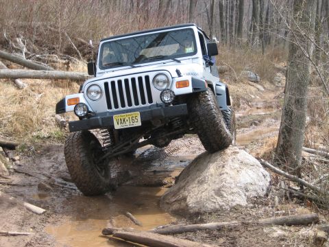 1990 Ford Bronco - This Jeep Wrangler TJ is shown off-roading on a challenging muddy trail, climbing over a large rock.