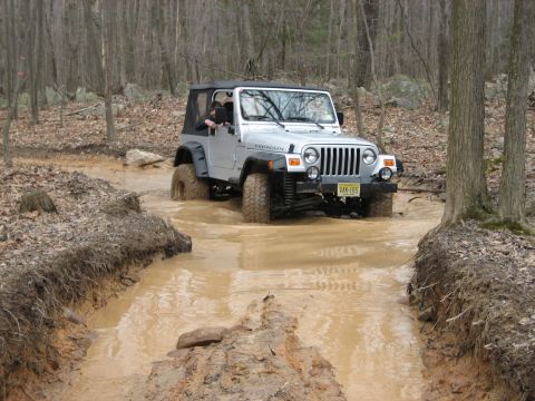 1990 Ford Bronco - A Jeep Wrangler navigates a challenging muddy path surrounded by trees.