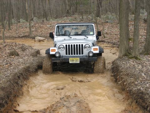 1990 Ford Bronco - A silver Jeep Wrangler navigates a muddy trail in a wooded area.