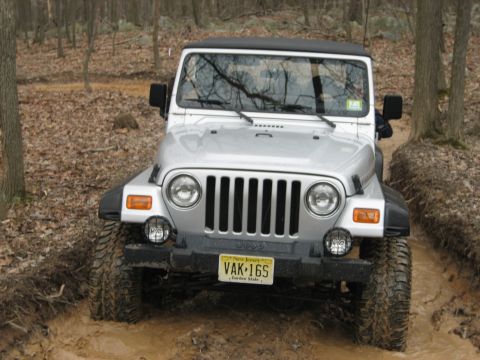1990 Ford Bronco - Silver Jeep Wrangler TJ navigating a muddy off-road trail in a wooded area.