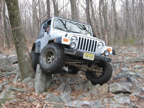 1990 Ford Bronco - A silver Jeep Wrangler TJ is shown off-roading over large rocks in a wooded area.