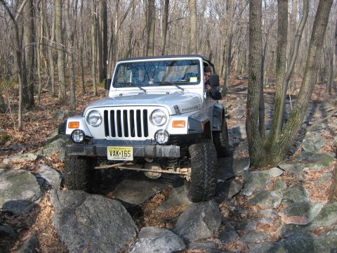 1990 Ford Bronco - Silver Jeep Wrangler tackling a challenging rocky path through a wooded area.