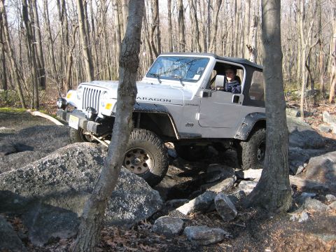 1990 Ford Bronco - Jeep Wrangler TJ off-roading through a wooded, rocky area. The vehicle appears to be a later model.