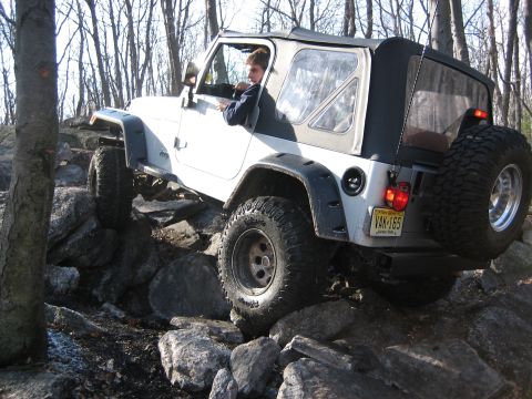1990 Ford Bronco - A white Jeep Wrangler tackles a challenging rocky trail with a driver at the wheel.