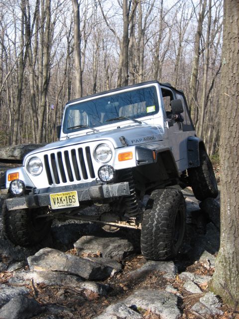 1990 Ford Bronco - A silver Jeep Wrangler TJ navigates a rocky trail with trees in the background.