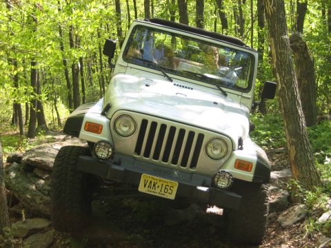 1990 Ford Bronco - A silver Jeep Wrangler navigates a challenging rocky path through a dense forest.