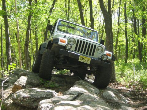 1990 Ford Bronco - A silver Jeep Wrangler navigates a rocky trail through a dense forest.
