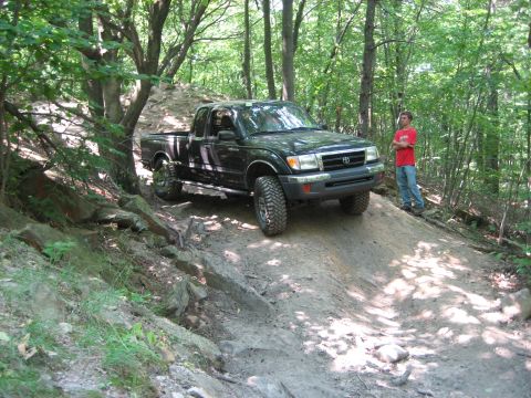 1990 Ford Bronco - A man watches as a black Toyota Tacoma navigates a challenging off-road trail through a forest.