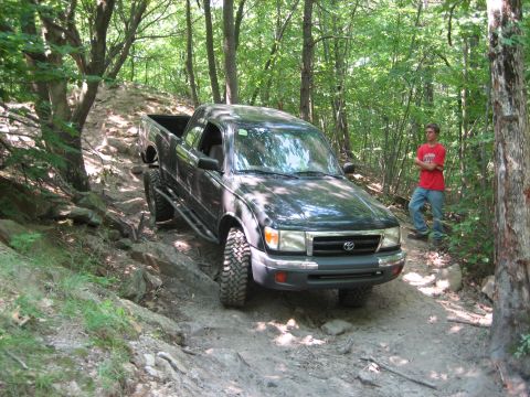 1990 Ford Bronco - A black Toyota Tacoma navigates a challenging off-road trail with a person standing nearby.